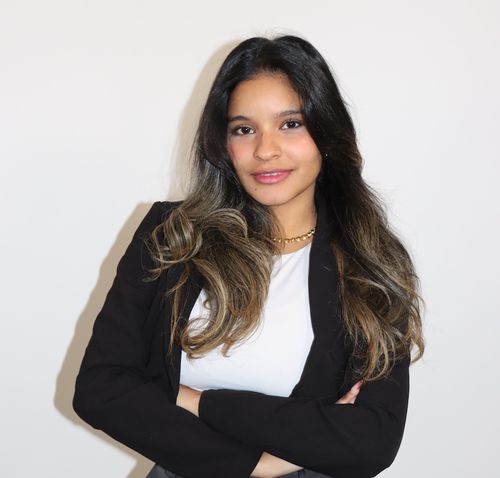 Smiling young woman with long dark hair wearing a black blazer and white top, standing with arms crossed against a white background.