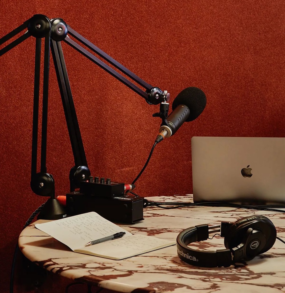Podcasting setup with a mounted microphone, headphones, a notebook with pen, and a laptop on a marble table against a red wall.