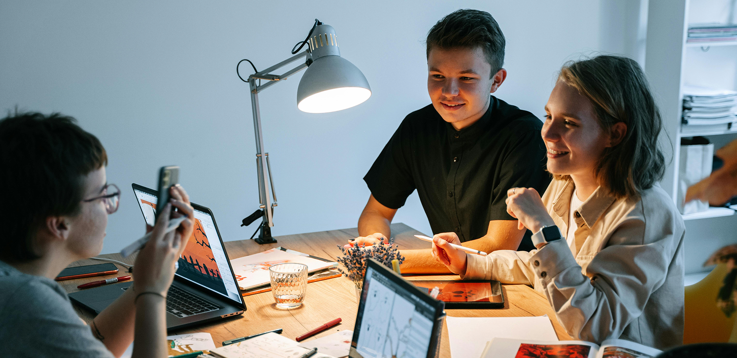 Three young adults collaborating around a wooden table with laptops, tablets, papers, and a desk lamp.
