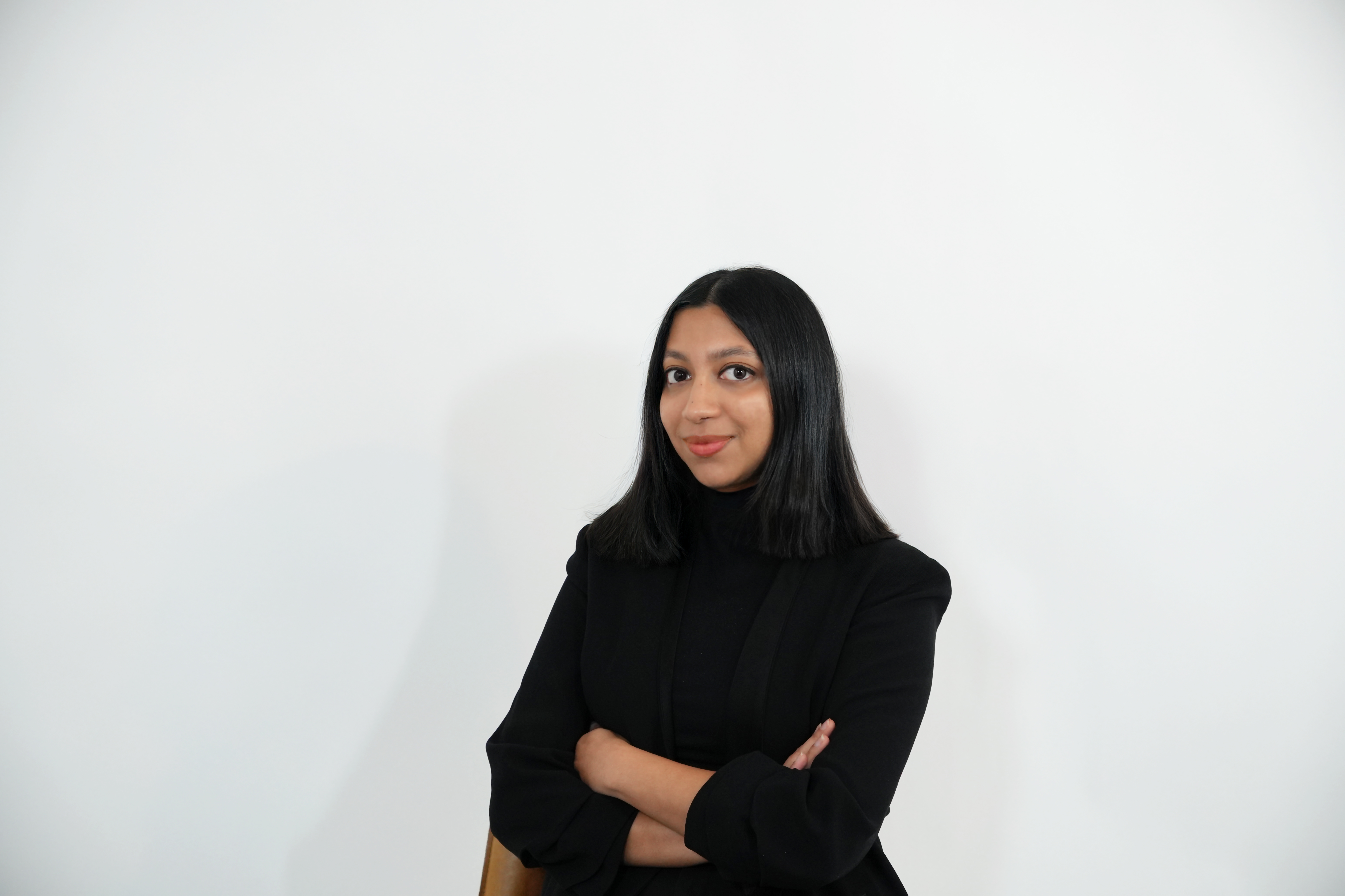 Woman with shoulder-length black hair wearing a black blazer and turtleneck, standing with arms crossed against a white background.