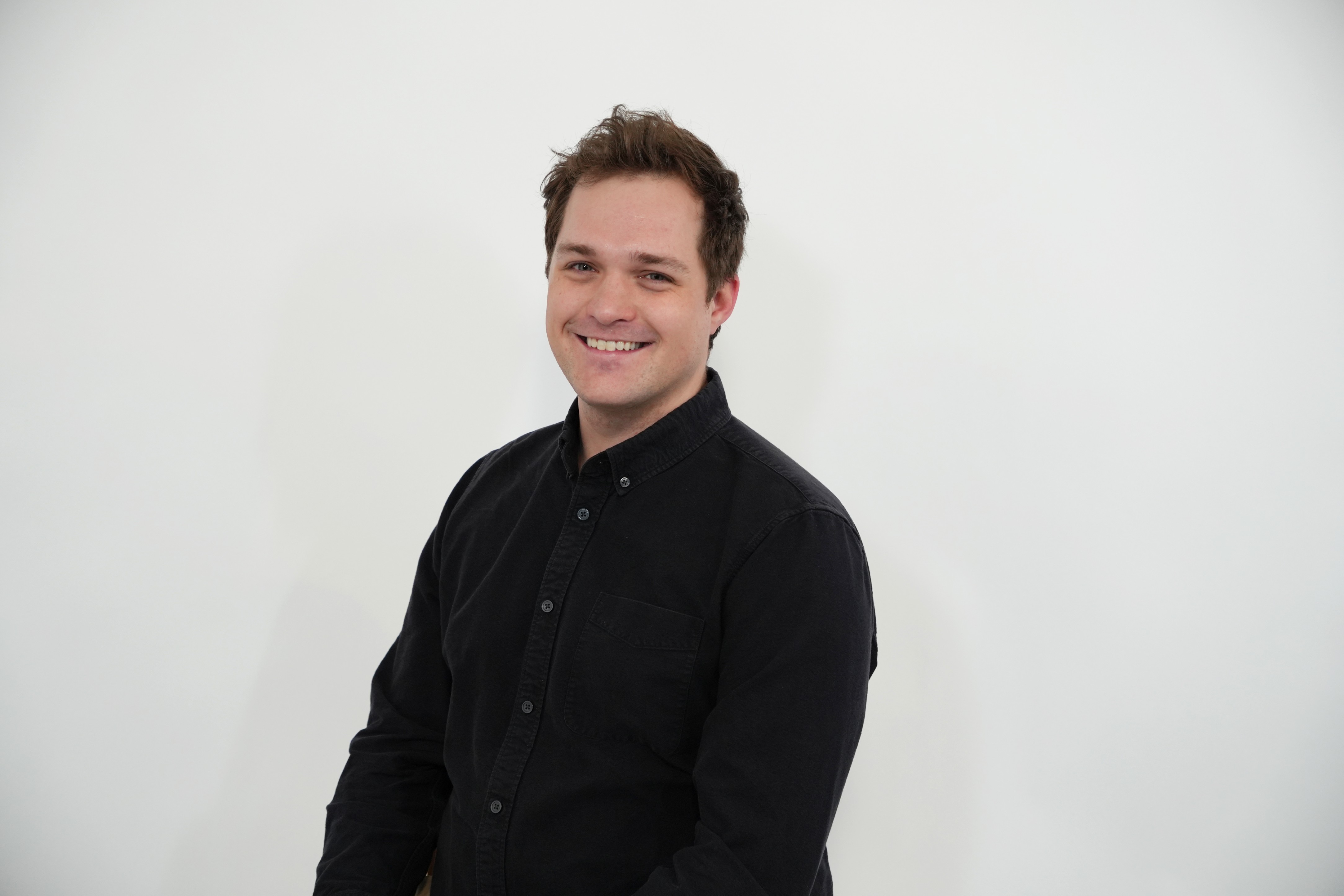 Smiling young man with short brown hair wearing a black button-up shirt against a plain white background.