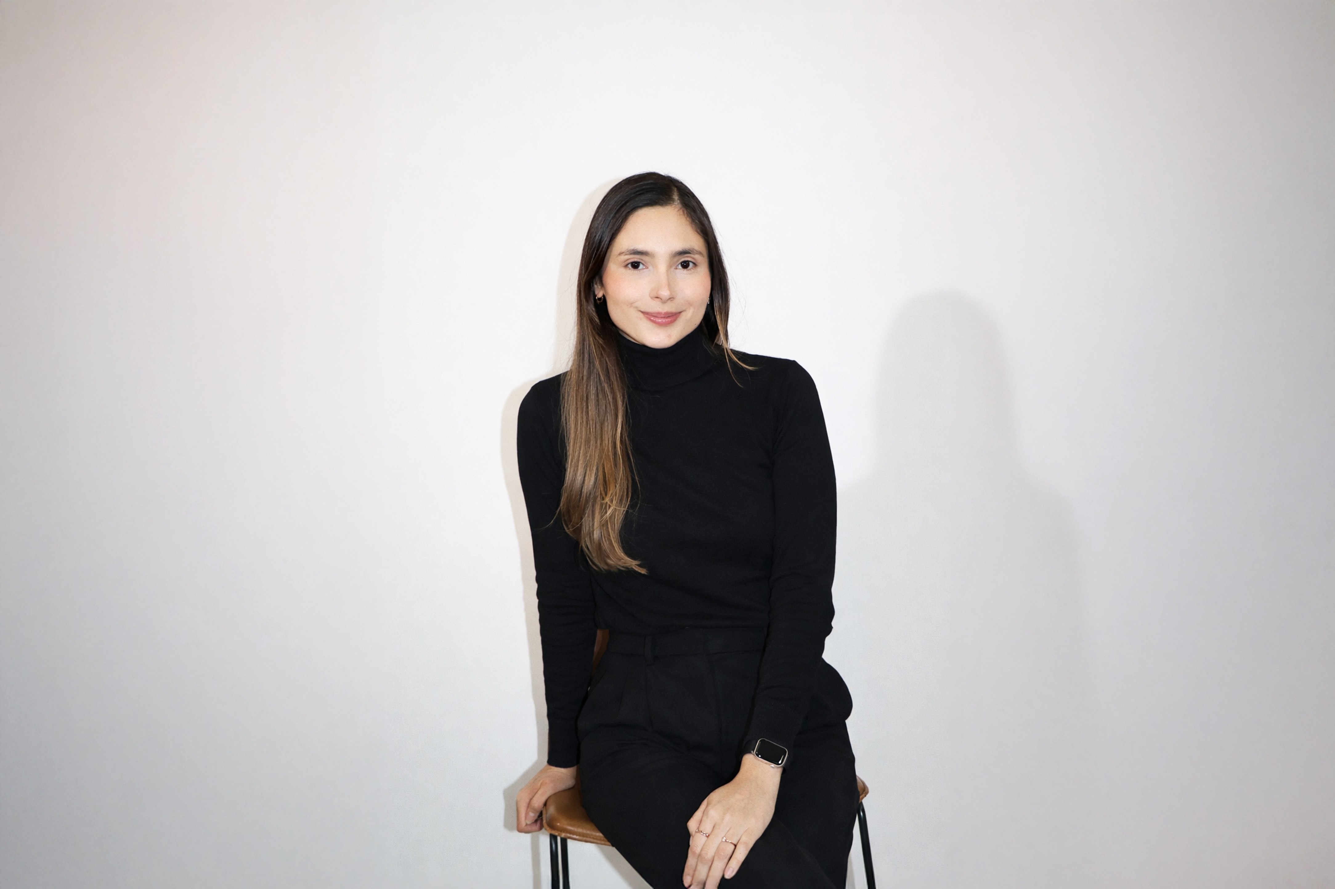 Young woman with long brown hair wearing a black turtleneck and pants, sitting on a chair against a plain white wall.
