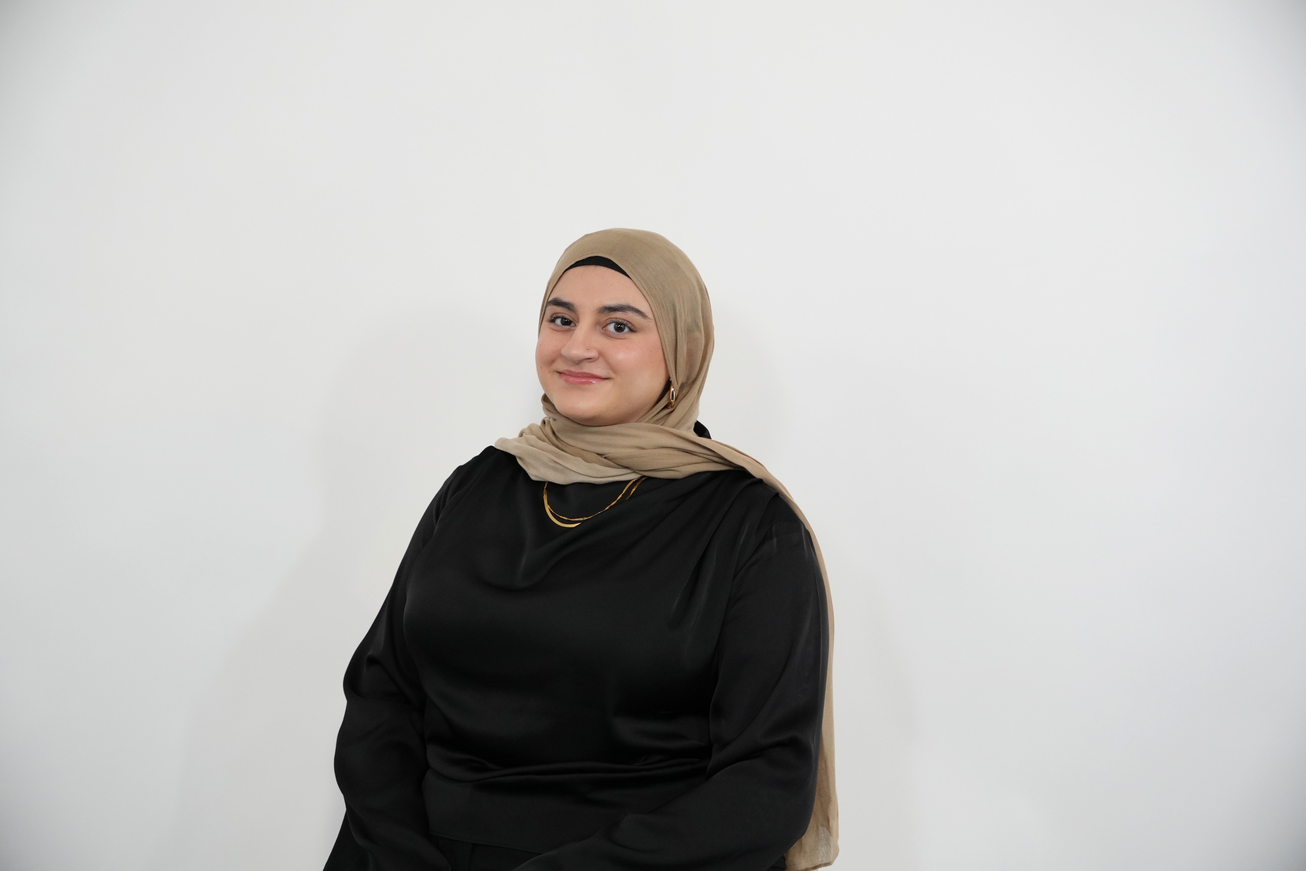 Woman wearing a beige hijab and black top smiling against a plain white background.