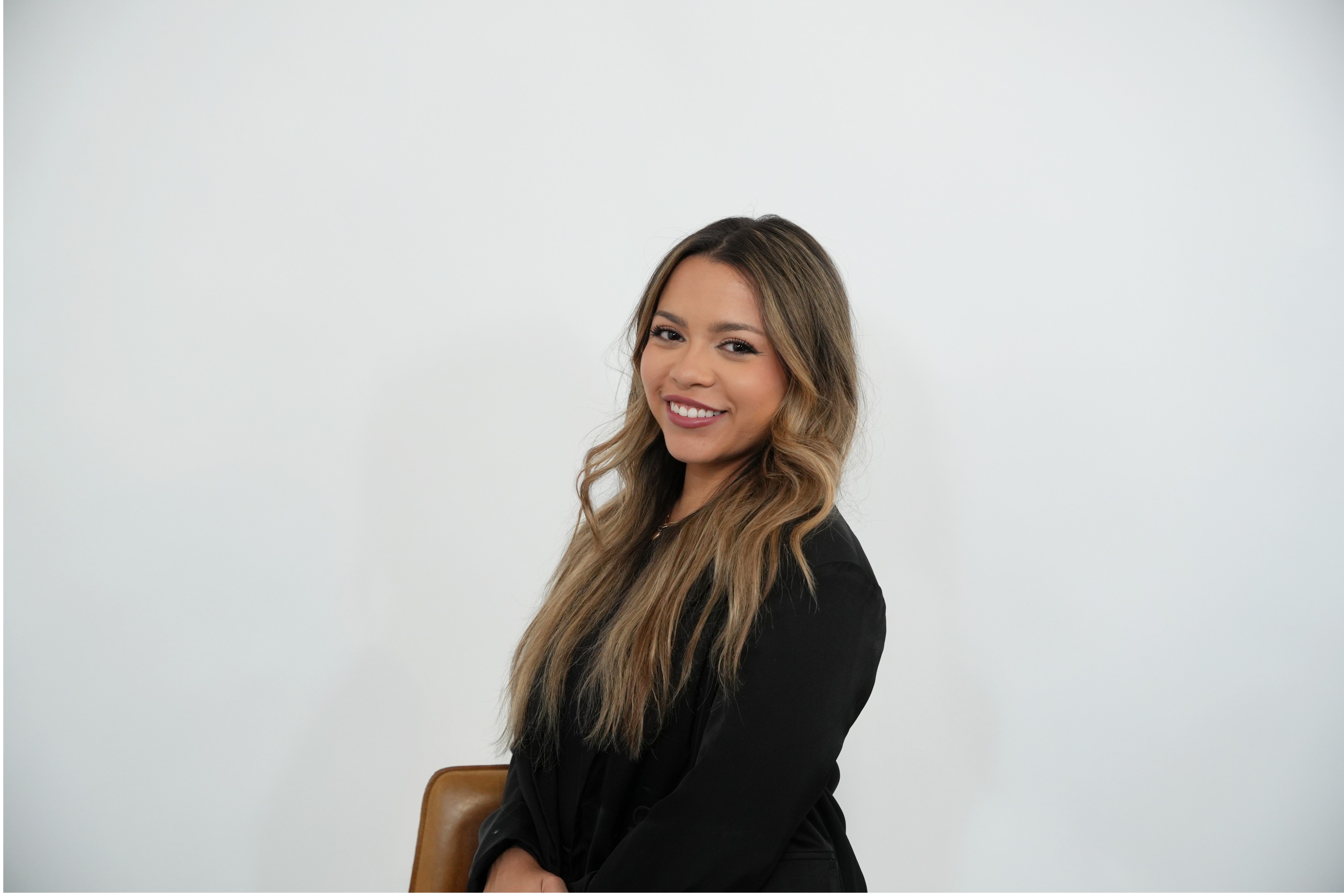 Smiling woman with long wavy hair wearing a black top seated on a brown chair against a plain white background.