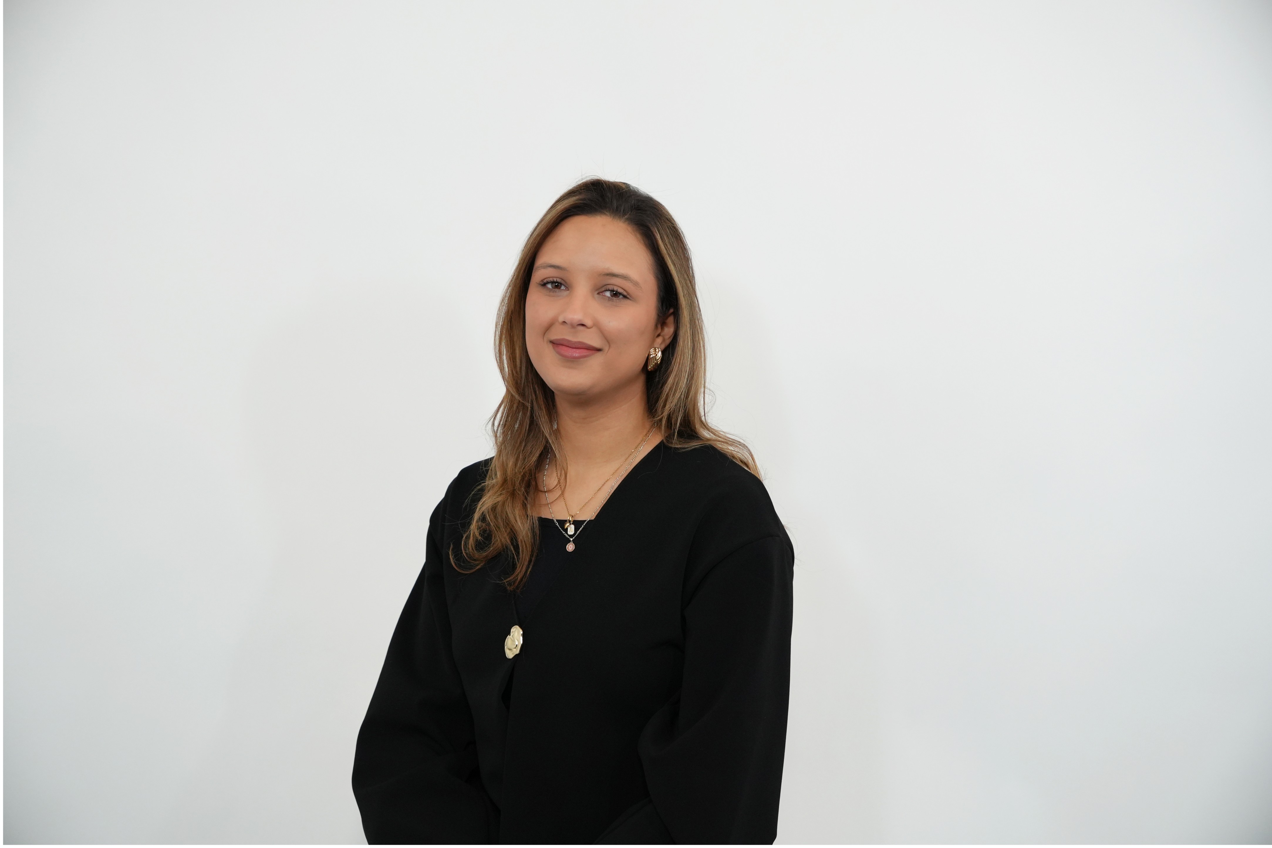 Smiling woman with light brown hair wearing a black top and gold jewelry, posed against a white background.