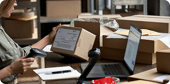 Person scanning a shipping label on a cardboard box with laptop and packing materials on the table.
