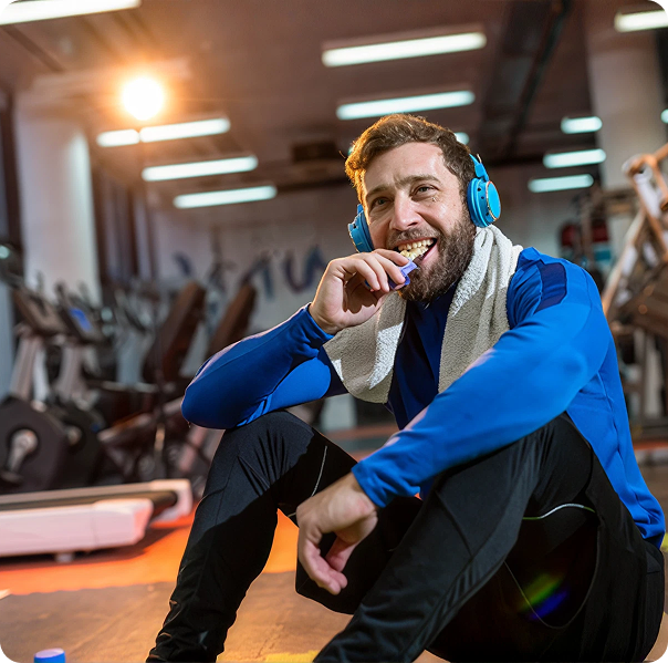 Smiling man wearing headphones and a towel eating a snack during a gym break.