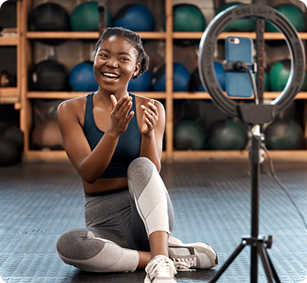 Smiling woman in workout clothes sitting on gym floor recording video with ring light and smartphone.