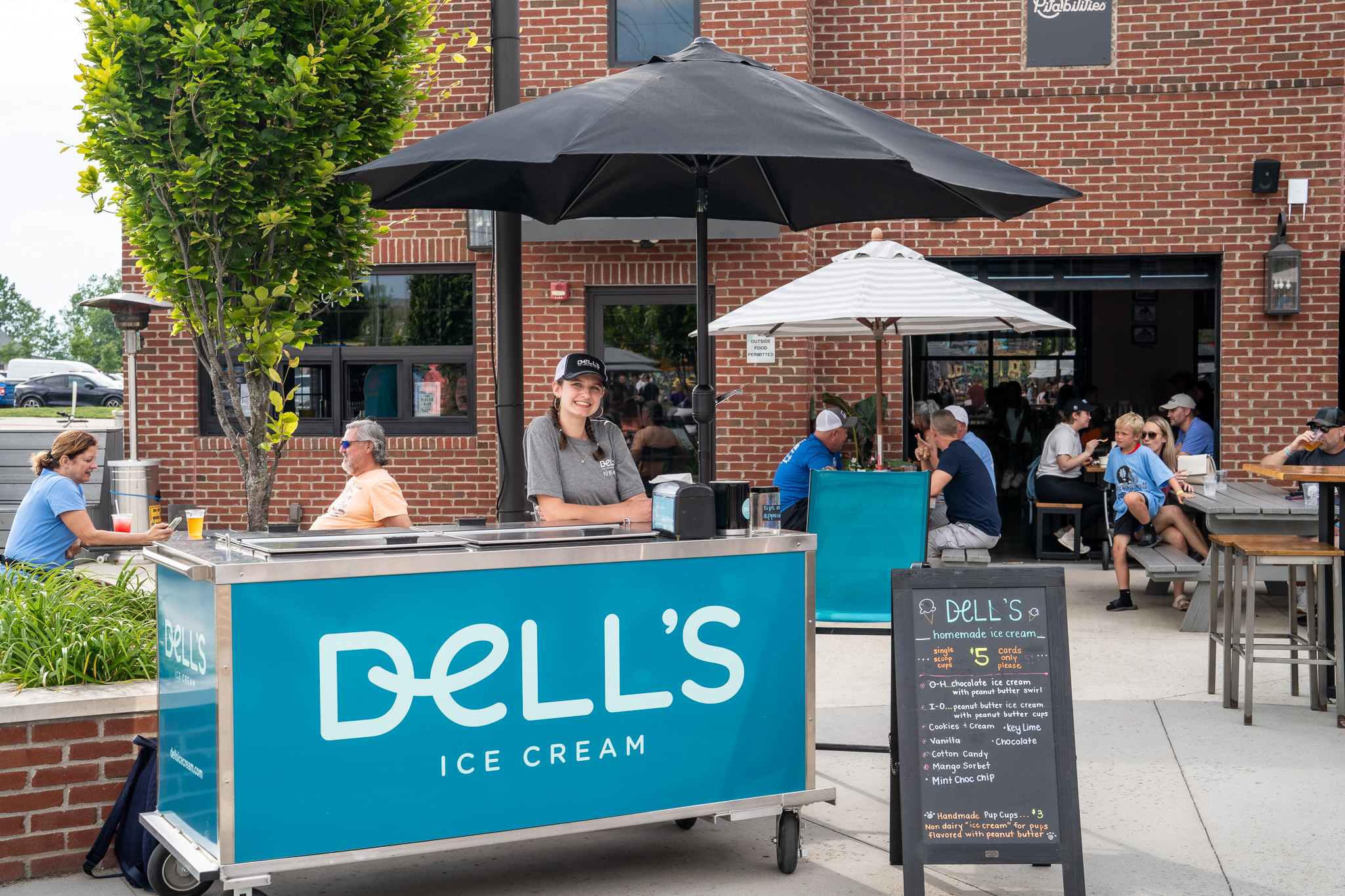 Outdoor Dell's Ice Cream cart with a smiling employee under a black umbrella and customers sitting at tables near a brick building.