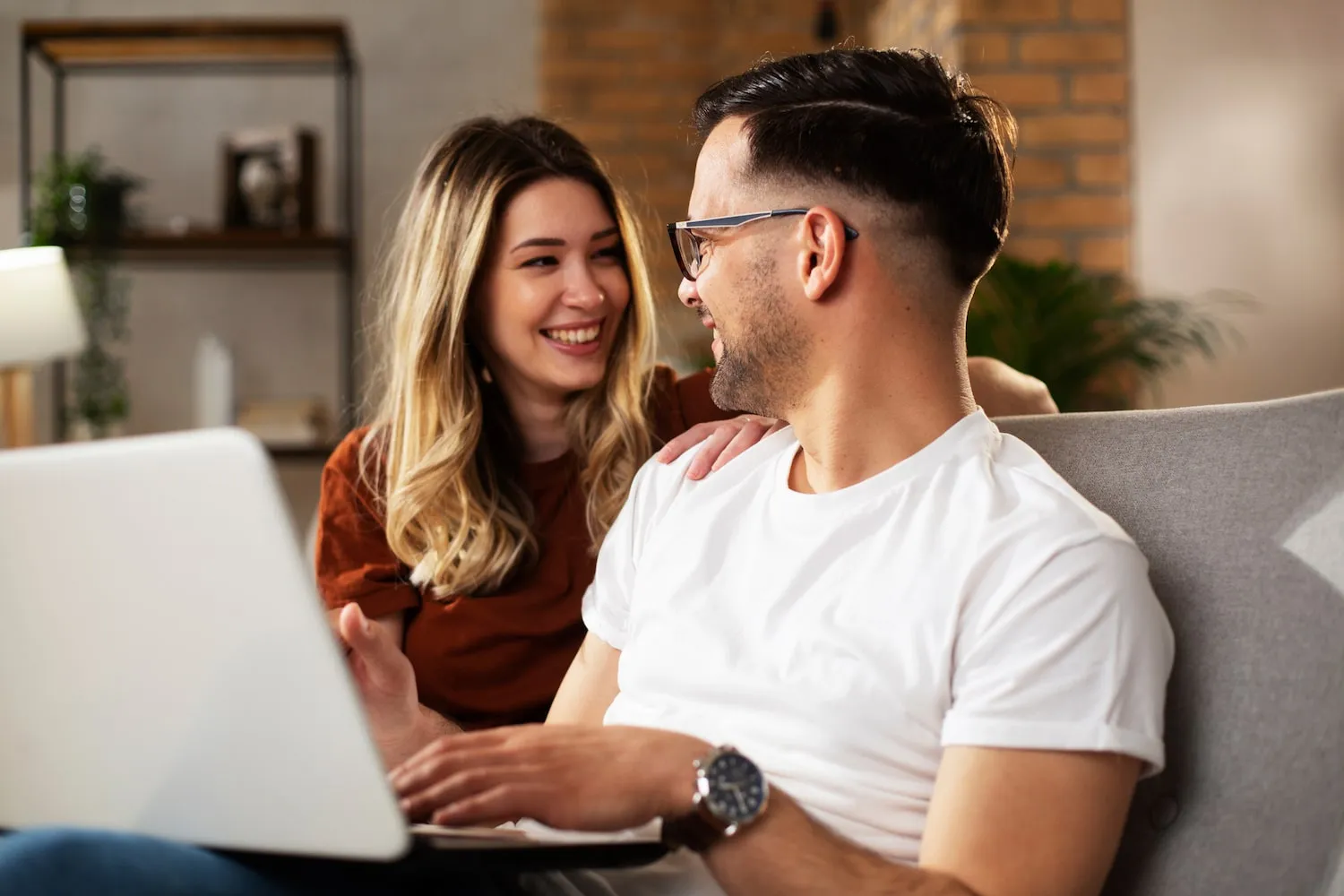 Smiling young couple sitting on a couch with a laptop, looking at each other.