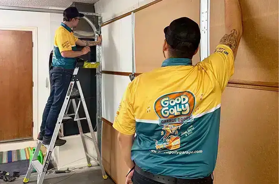 Two technicians in yellow and teal Good Golly Garage Doors shirts installing or repairing a garage door inside a residential space, one standing on a ladder.
