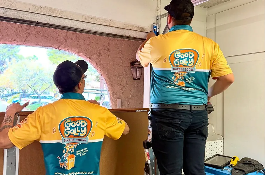 Two workers in yellow and teal Good Golly Garage Doors shirts installing or repairing a garage door inside a garage.