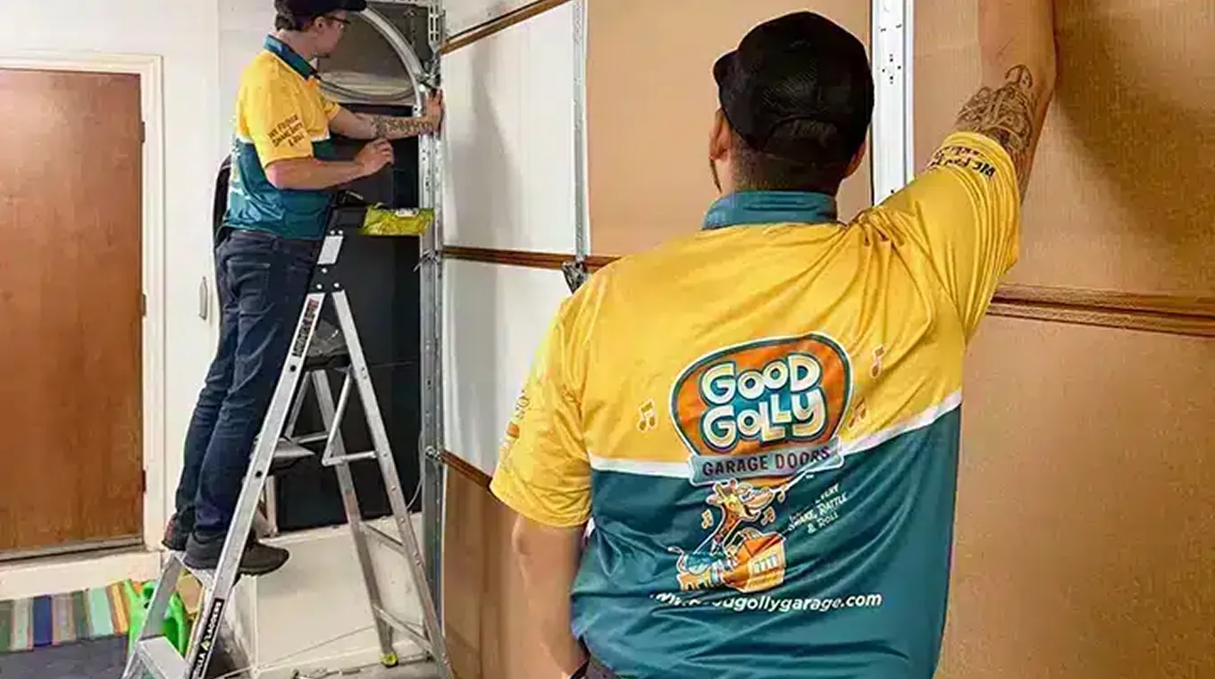 Two technicians in Good Golly Garage Doors uniforms installing or repairing a garage door, one on a ladder and one standing.