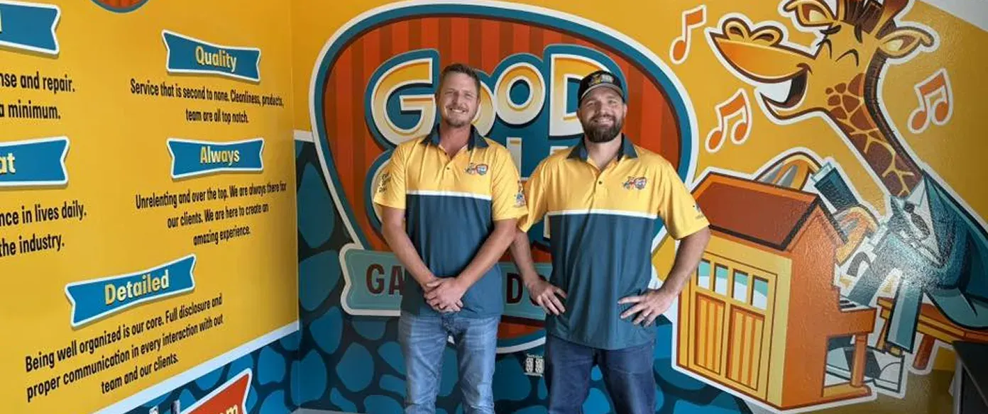 Two men wearing matching yellow and blue polo shirts stand smiling in front of a colorful wall mural with text and a cartoon giraffe holding a house model.