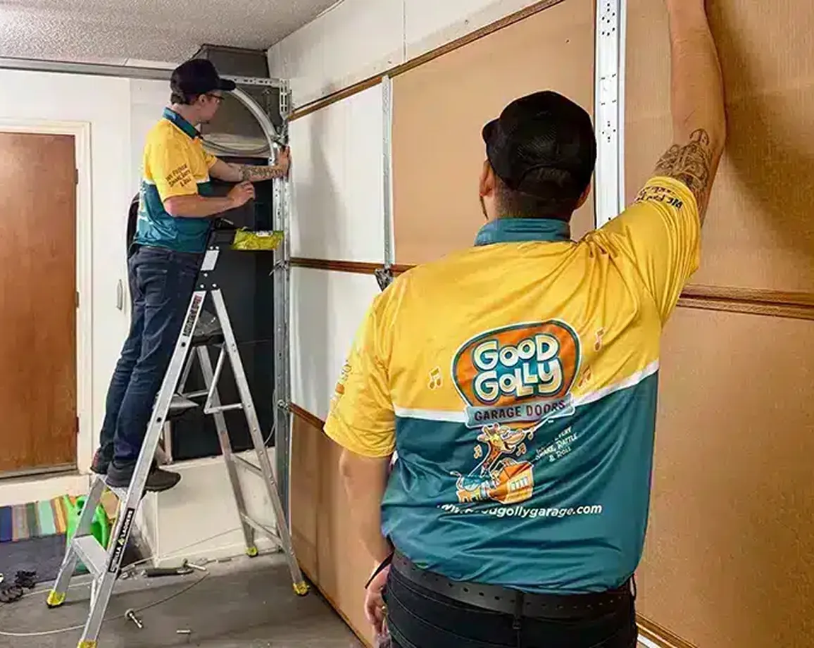 Two workers wearing yellow and green Good Golly Garage Doors shirts, installing or repairing a garage door, one standing on a ladder.