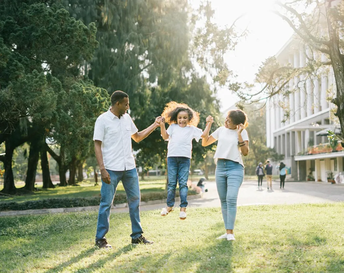 A man and a woman holding a smiling child’s hands and swinging her in a sunny park with trees and a building in the background.