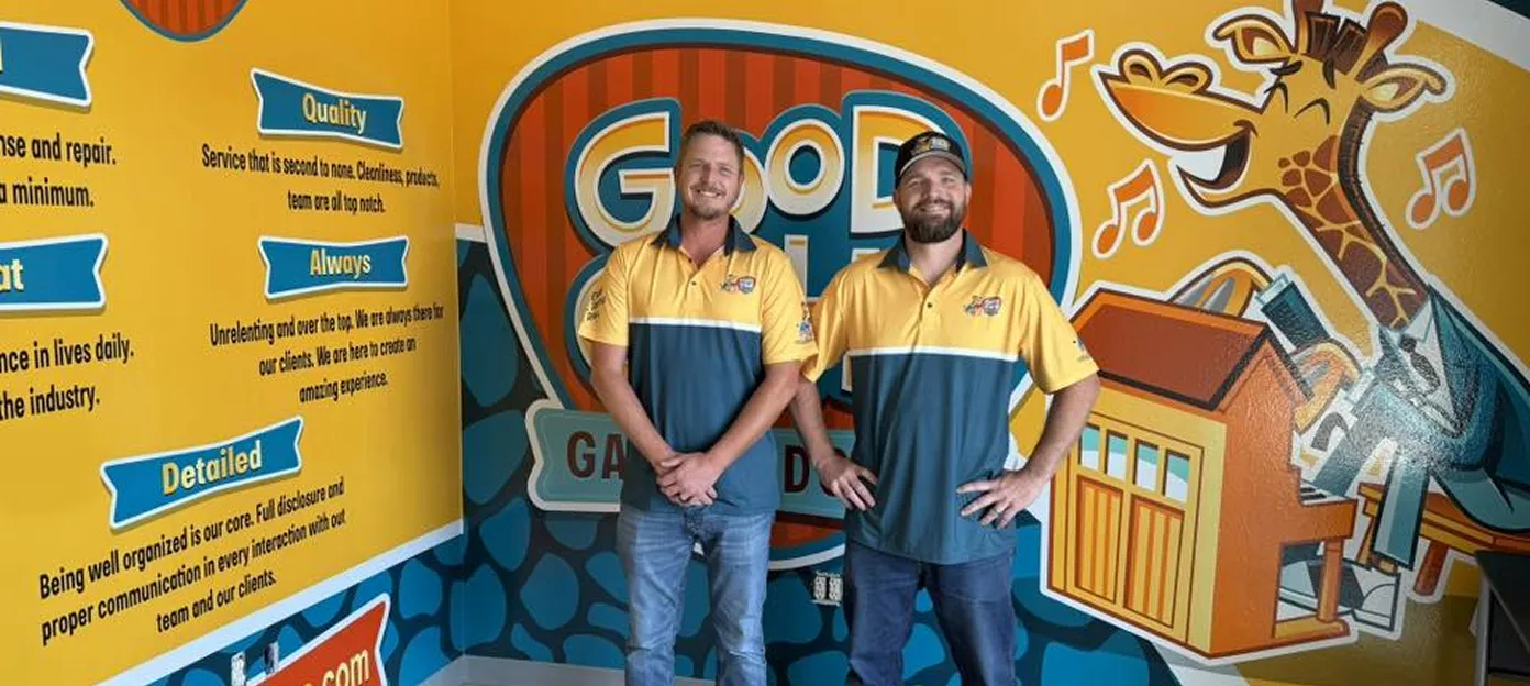 Two men wearing yellow and blue polo shirts stand smiling in front of a colorful wall mural with a giraffe and the words 'Good Guys Garage Doors'.