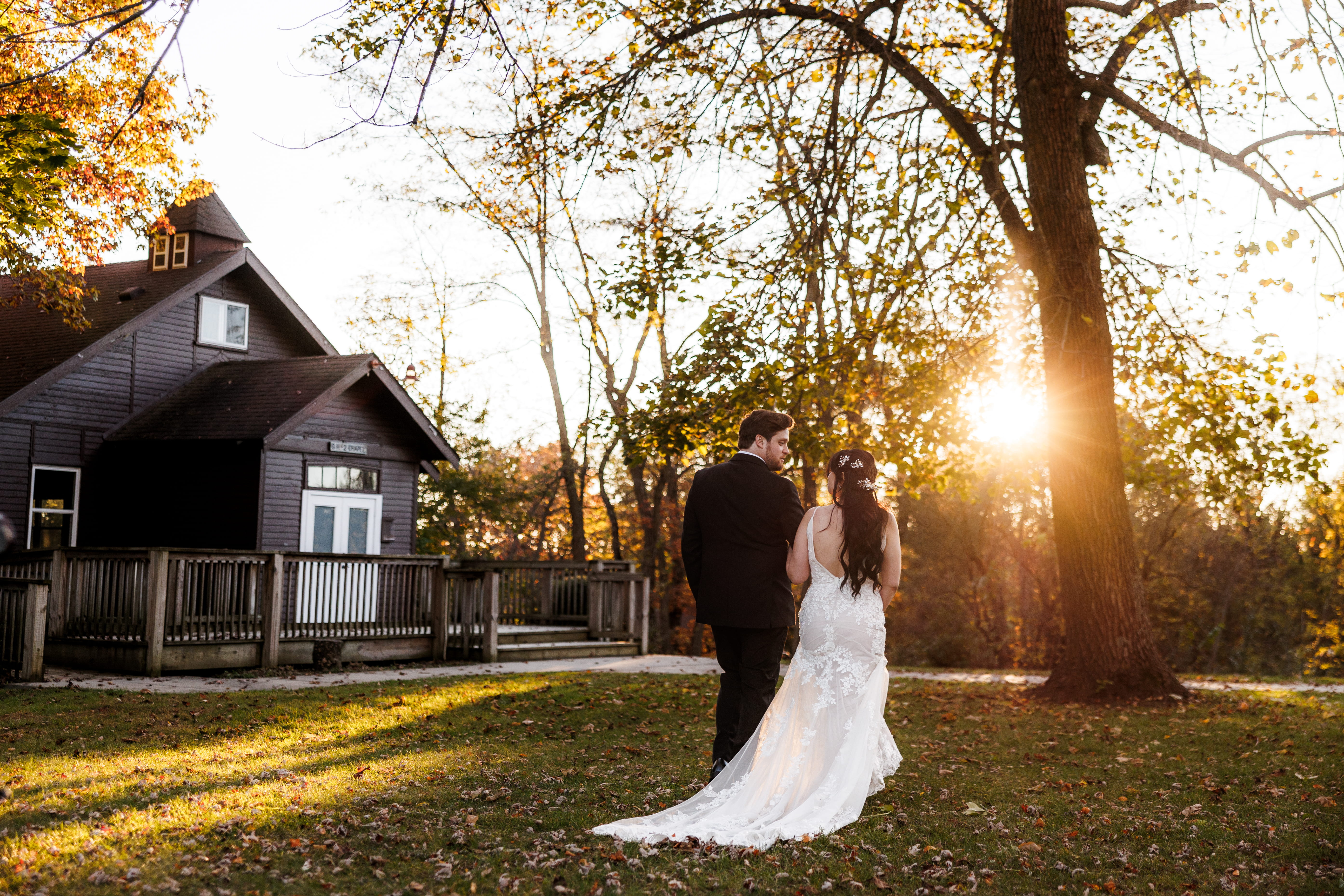 Man in a dark suit standing in tall dry grass with leafless trees and a streetlamp in the background at sunset.