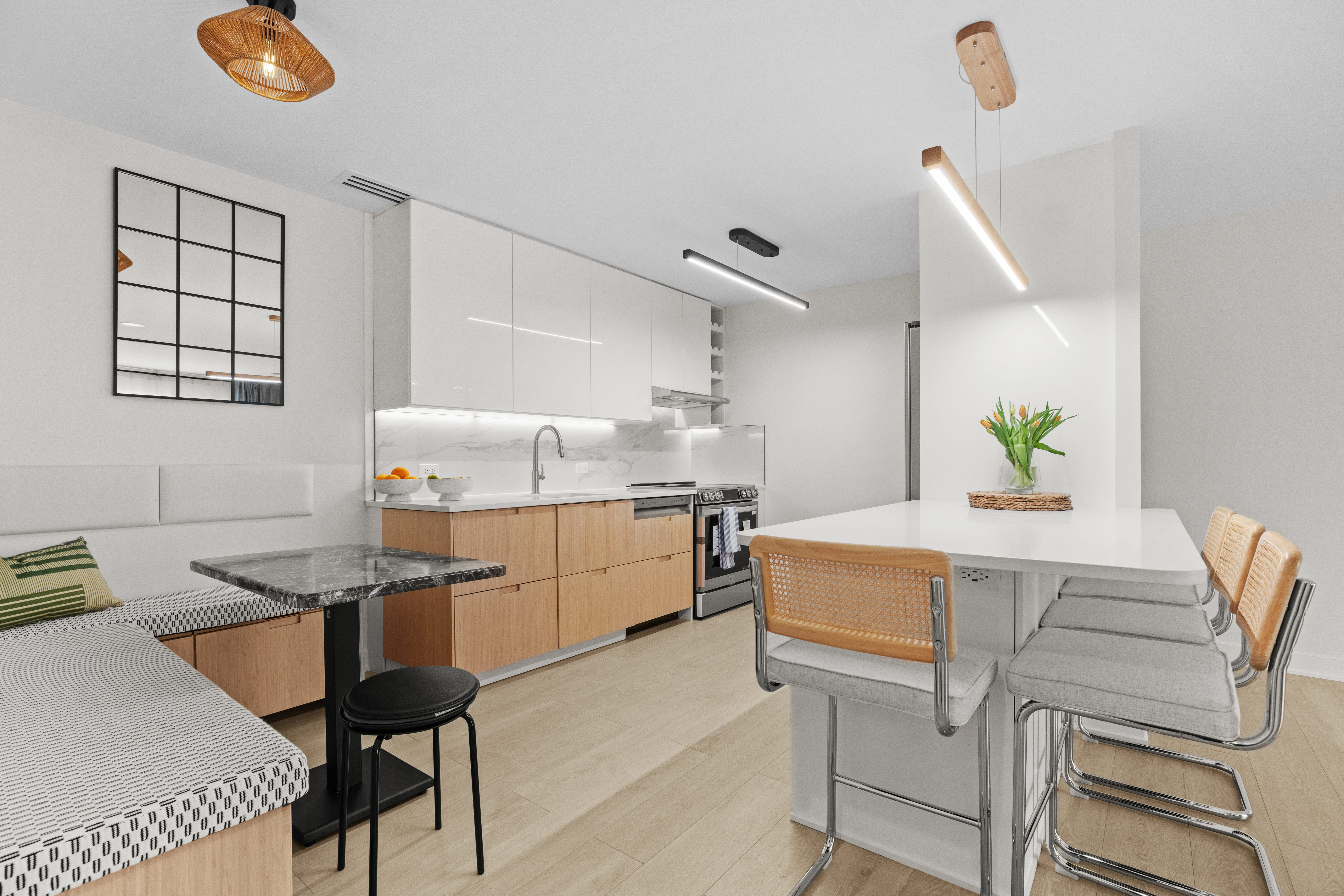 Modern kitchen with white and wood cabinets, a black marble small table with bench and stool, and a white island with four light-colored cushioned chairs and a vase of yellow tulips.