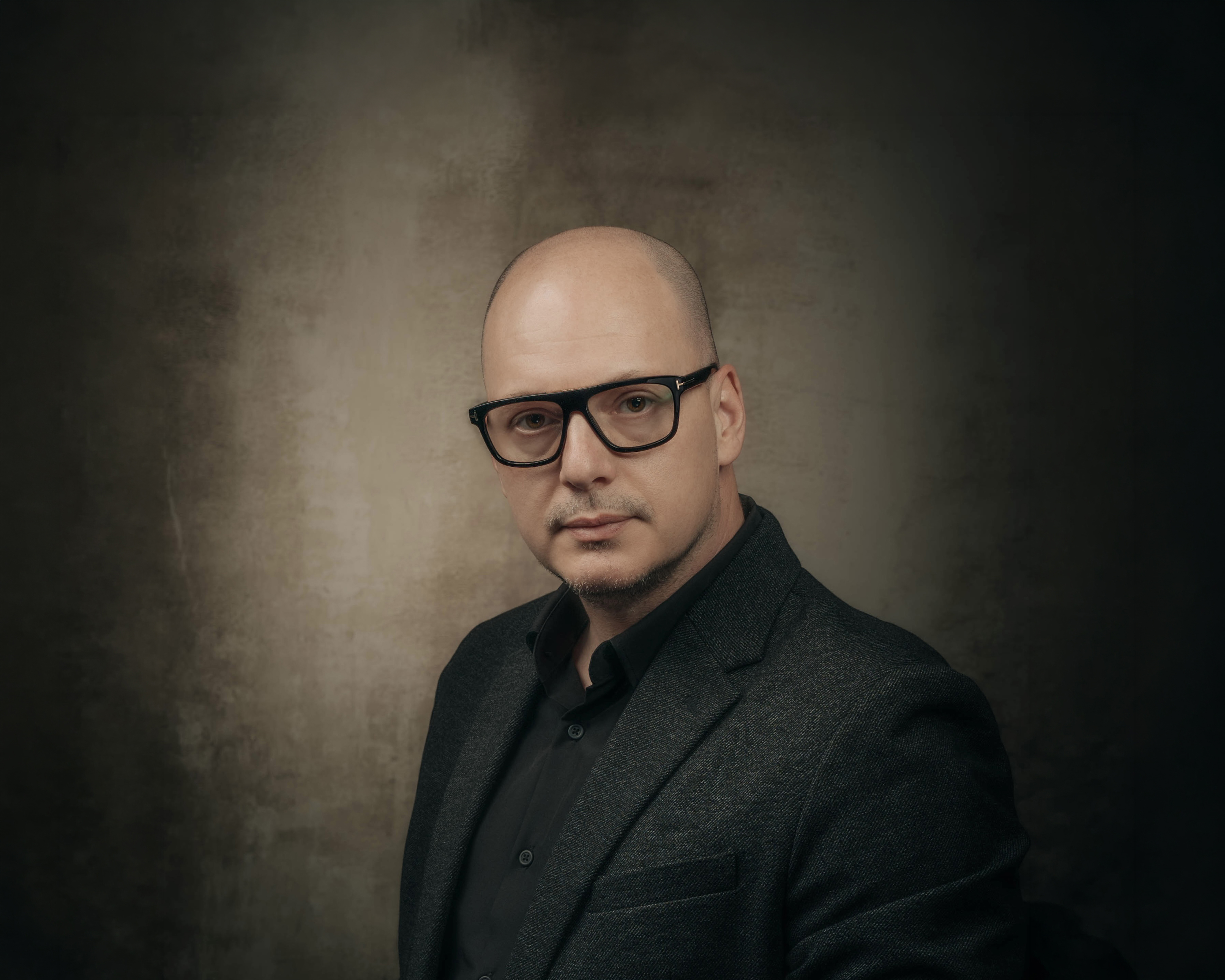 Bald man wearing black glasses and a dark suit jacket against a muted textured background.