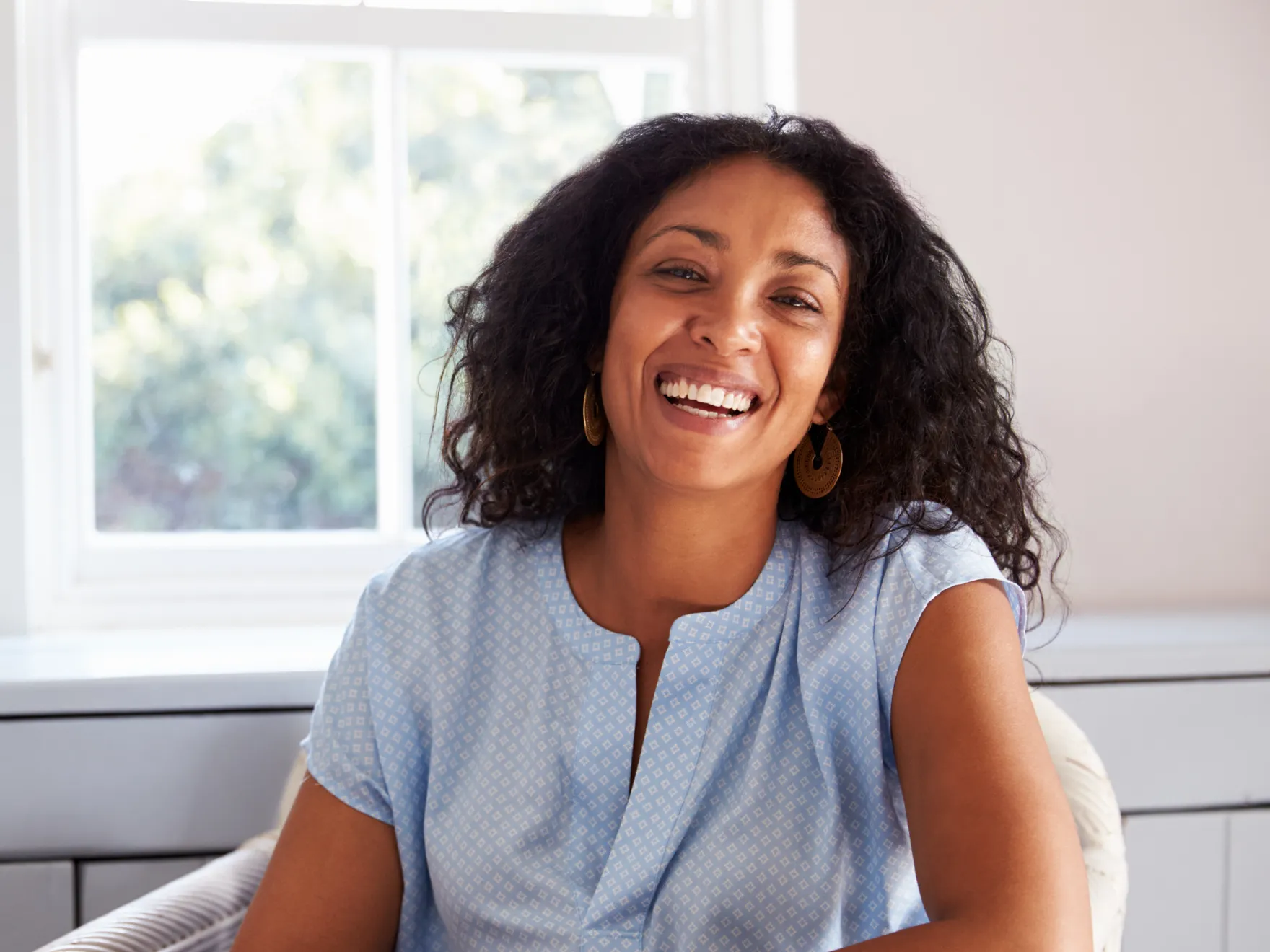 Woman smiling warmly, sitting indoors near a window.
