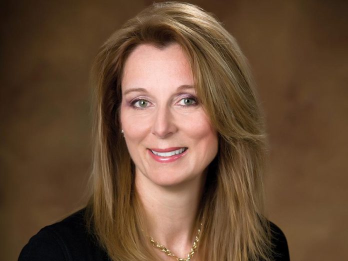 A woman with long light brown hair smiles, wearing a black top and gold necklace.