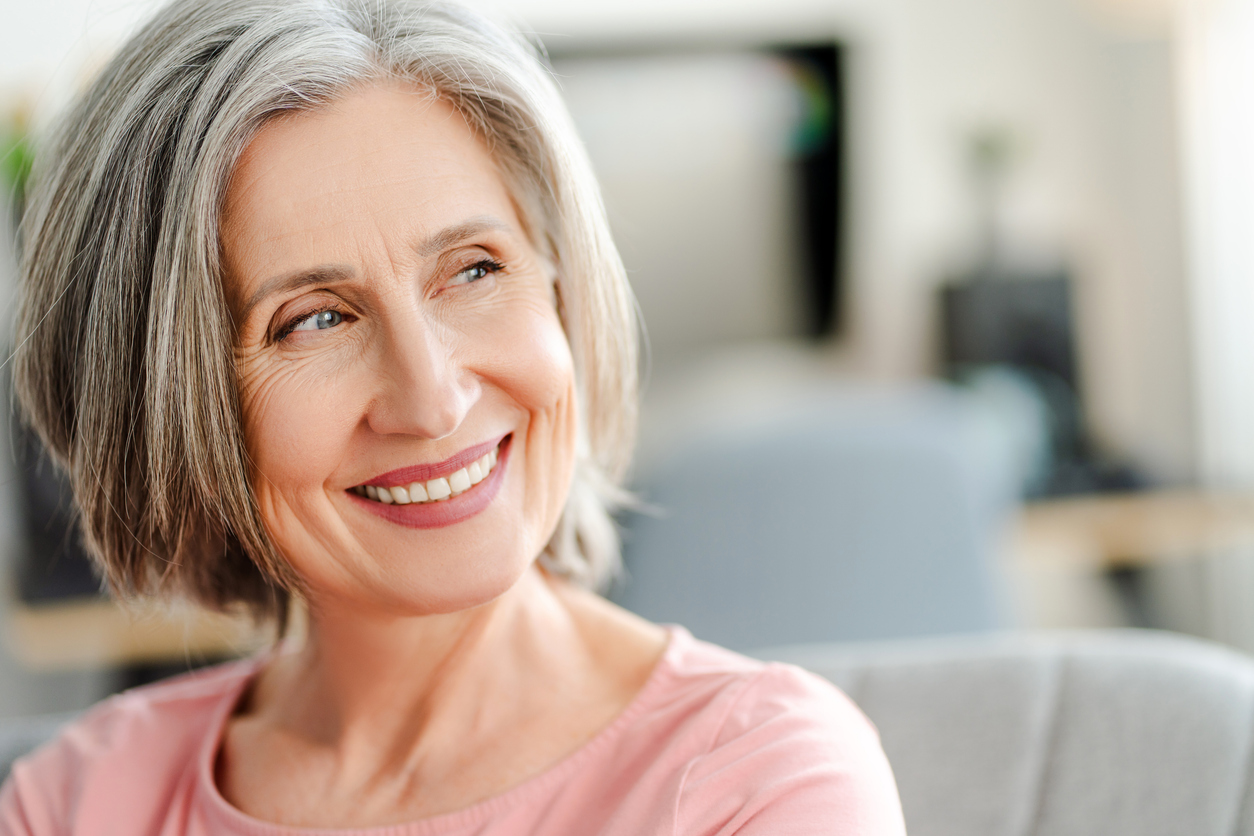 An older woman with gray hair smiles while sitting indoors, wearing a pink top.
