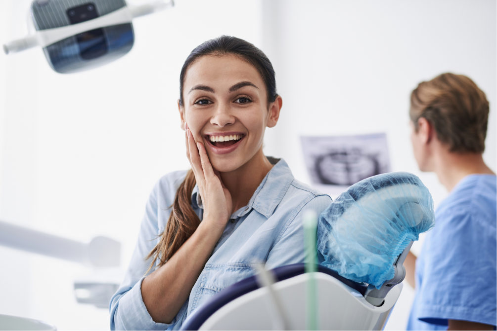 A smiling woman sits in a dental chair, holding her cheek, with dentist equipment nearby.