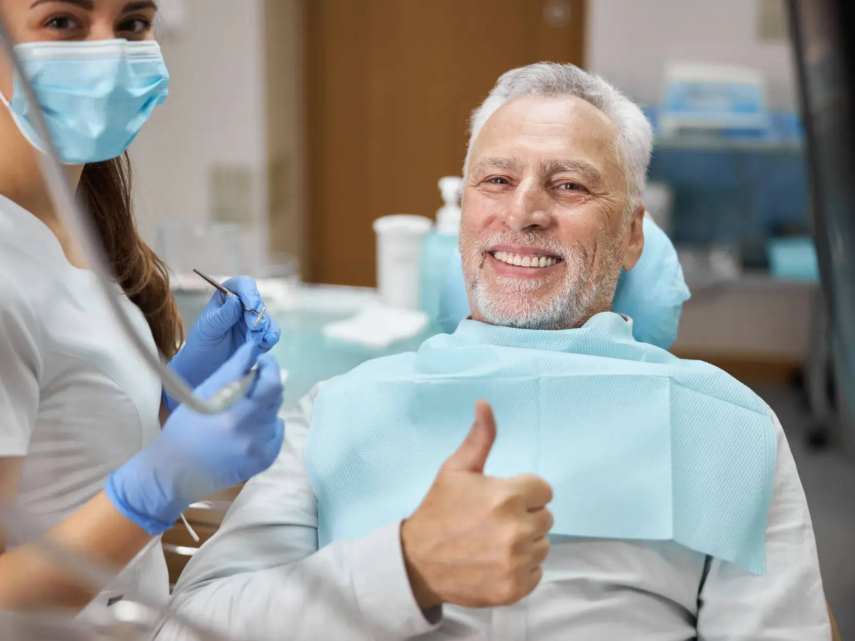 Dentist wearing a mask and gloves examines a smiling patient with sunglasses in a dental chair.