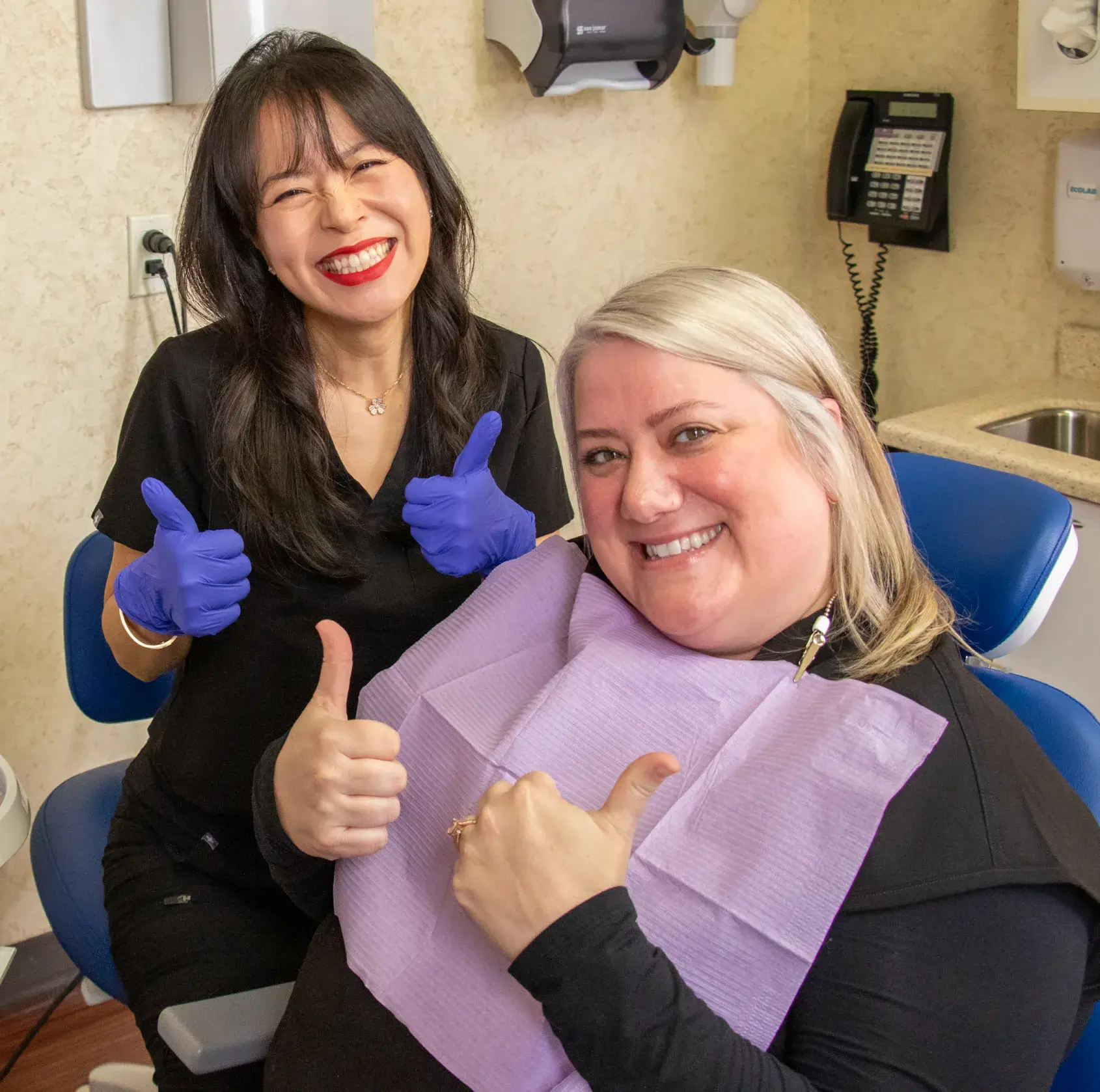 Two women in black uniforms smiling at each other, one seated in a dental chair.