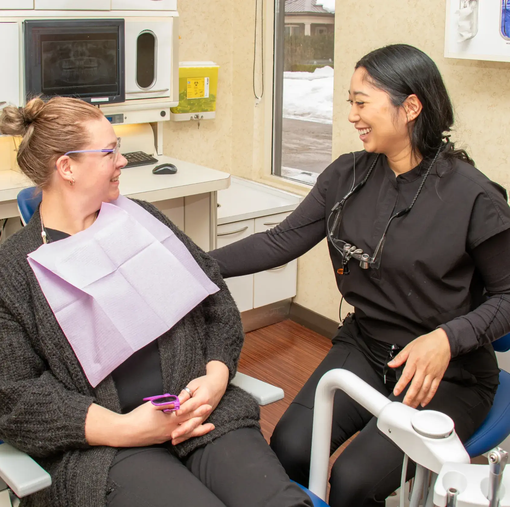 A woman in a black shirt stands smiling in a dental office with equipment around her.