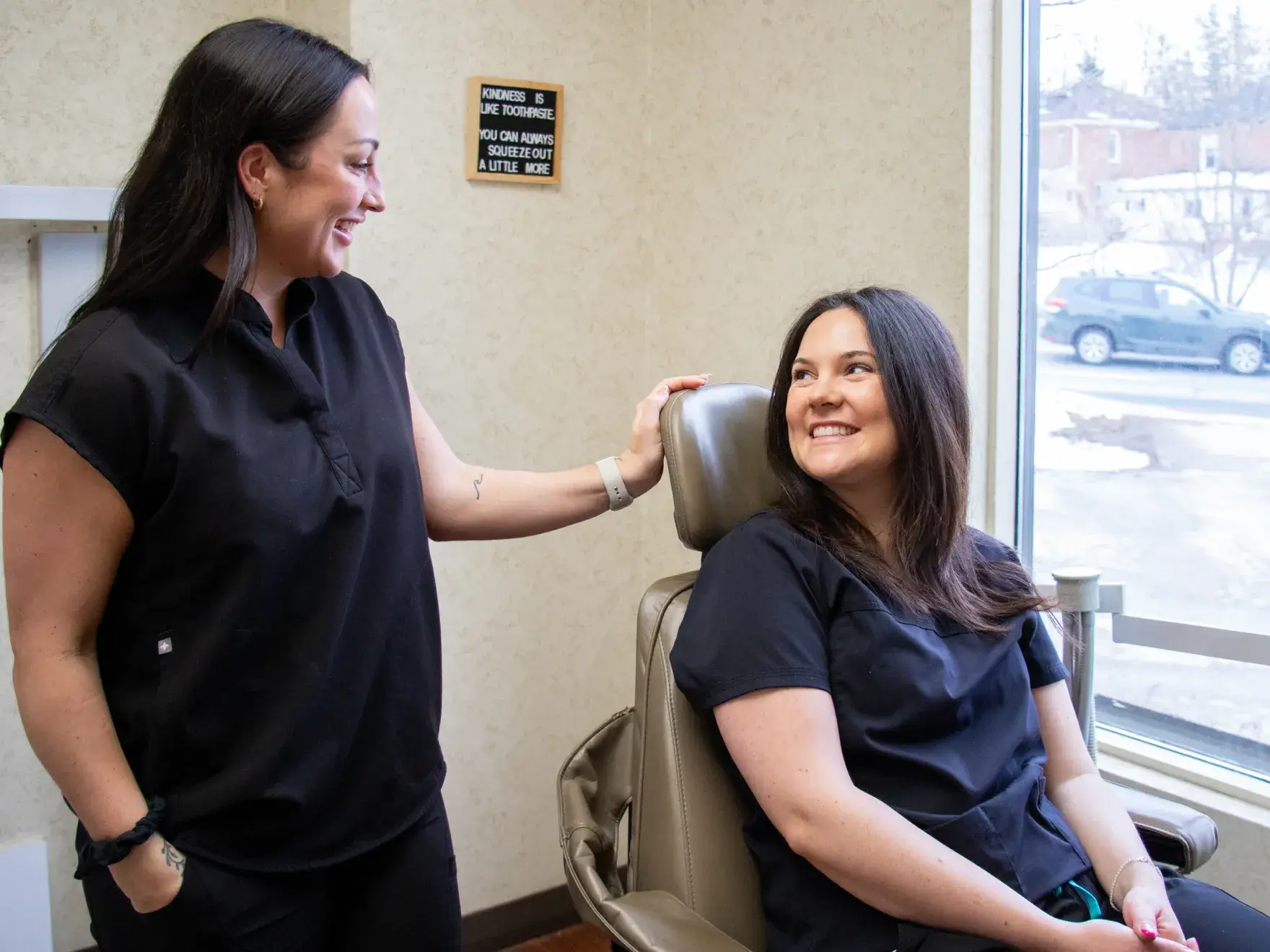A woman in dental attire smiles while standing in a dental office with arms crossed.