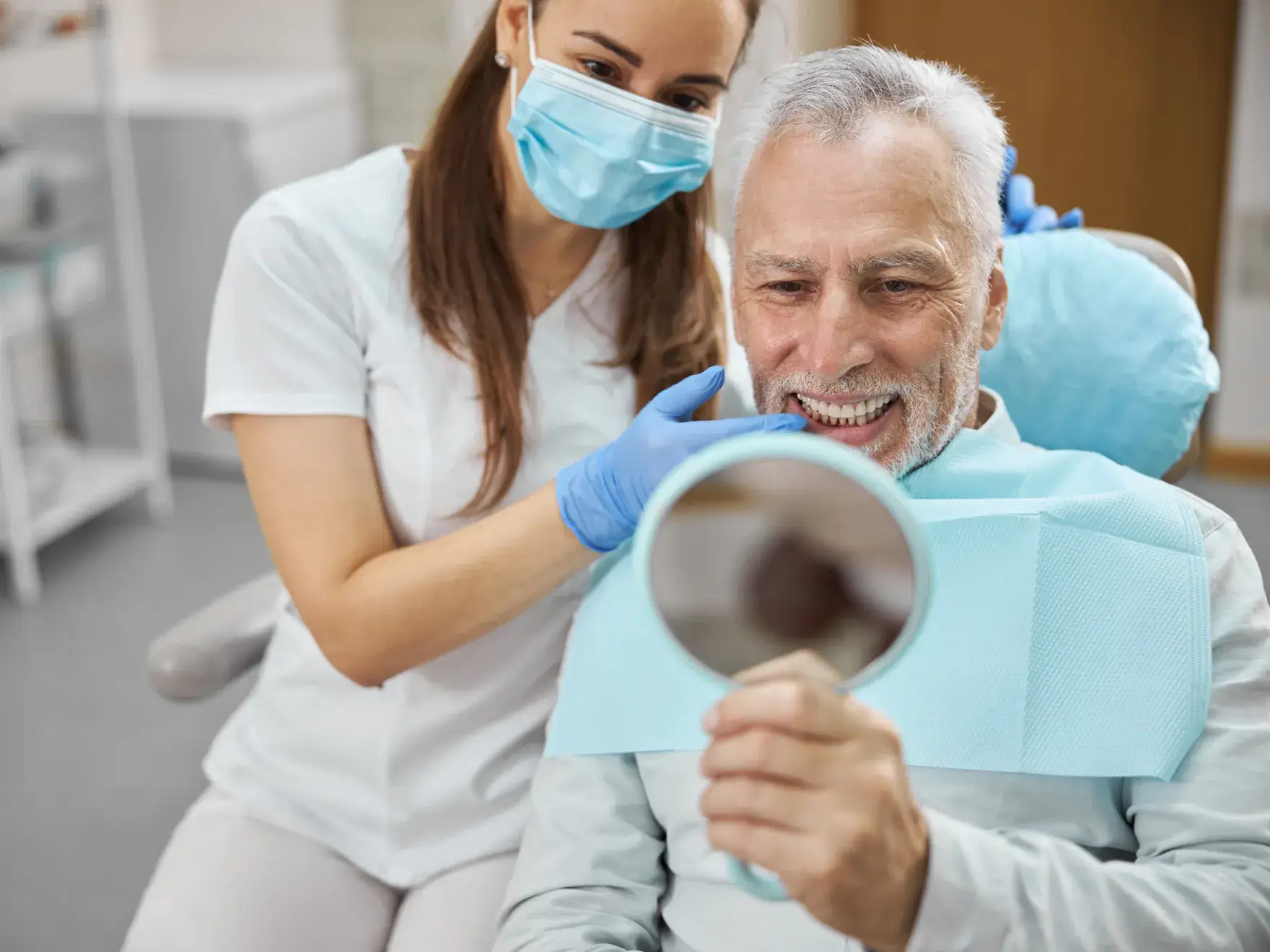 A dentist shows an elderly man his smile in a handheld mirror during a dental check-up.