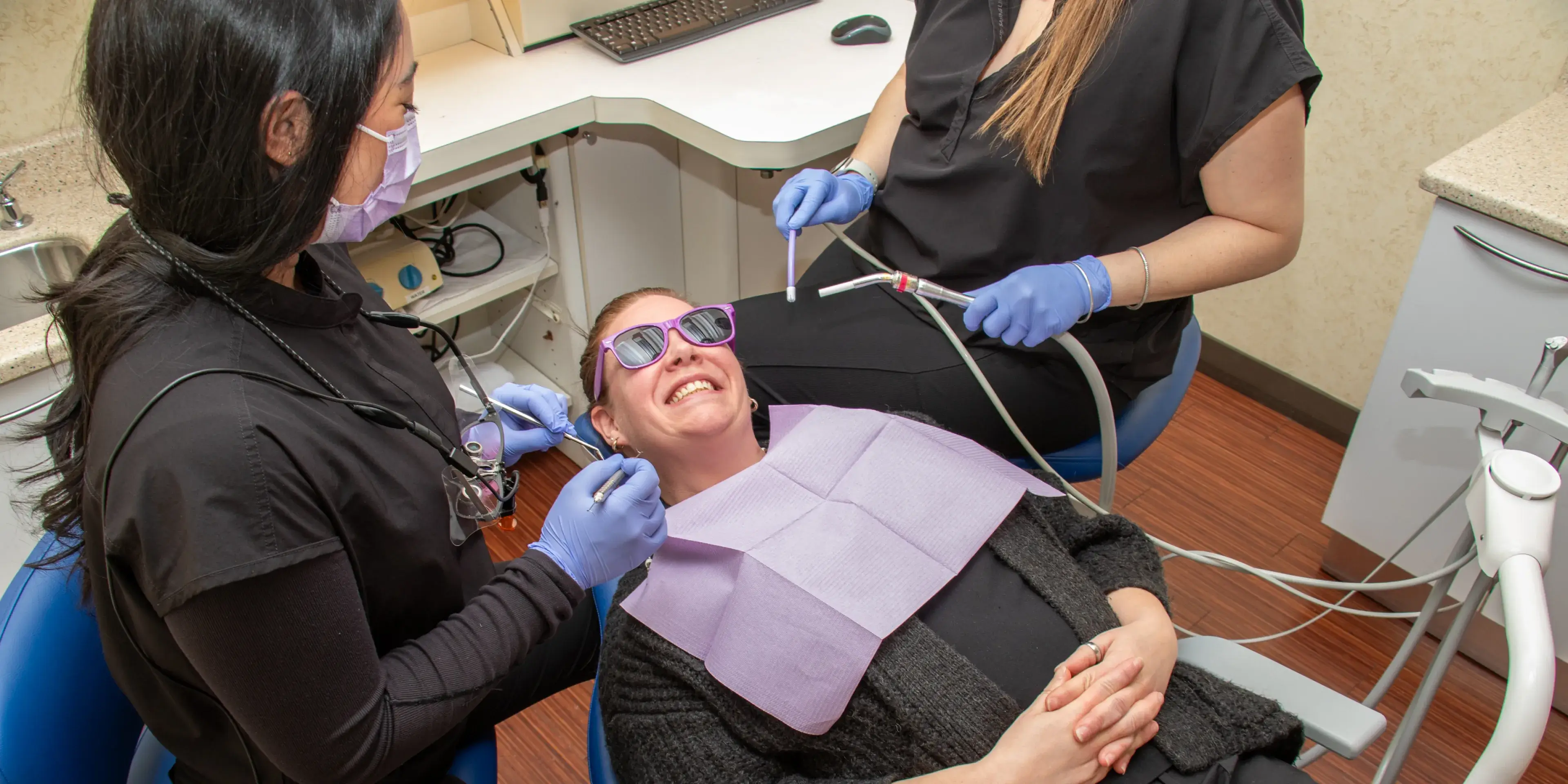 A person in sunglasses sits in a dental chair, while two dental assistants work on their teeth.