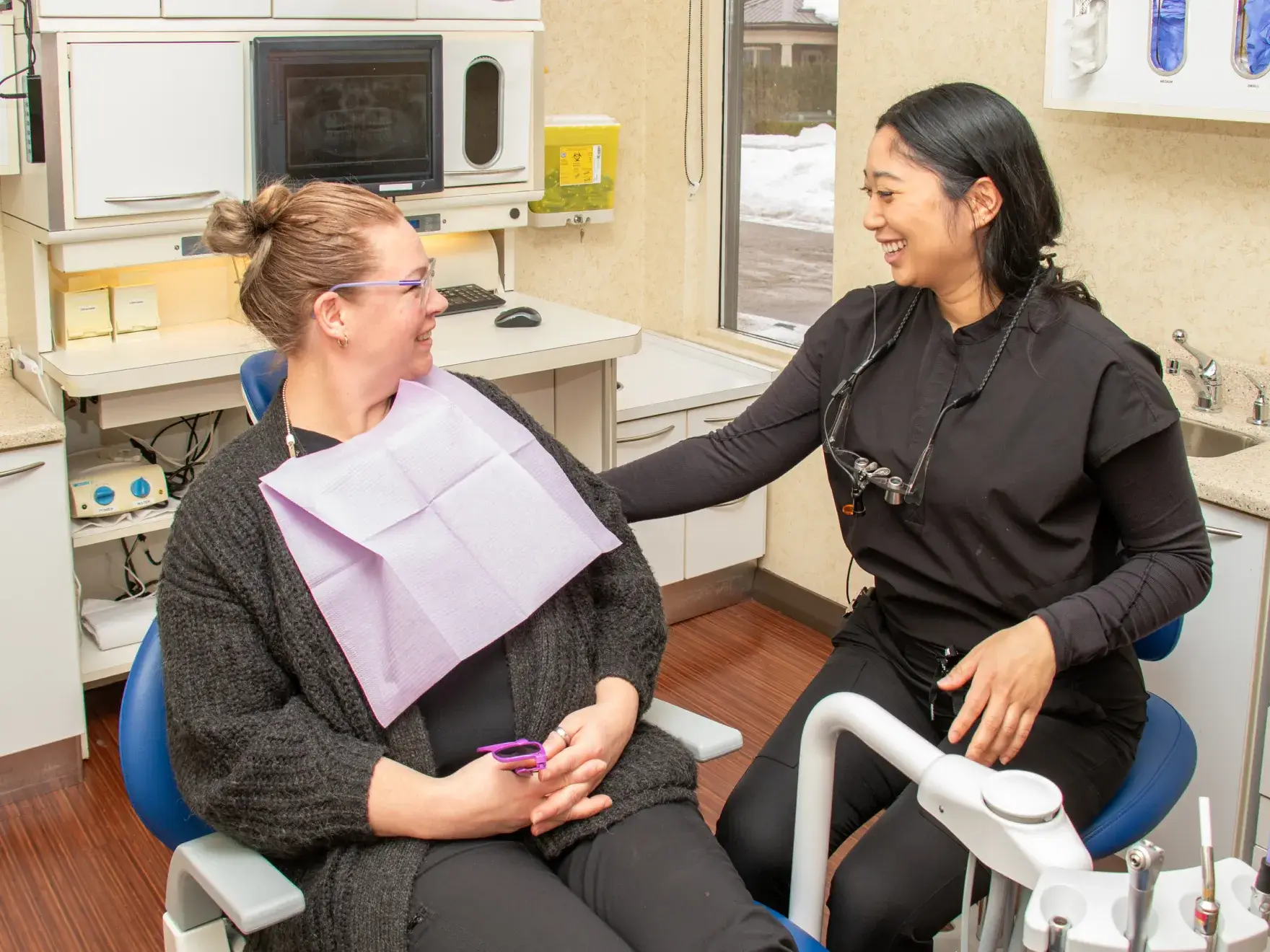 Two women in a dental office wearing black scrubs, one looking at a screen and another seated.