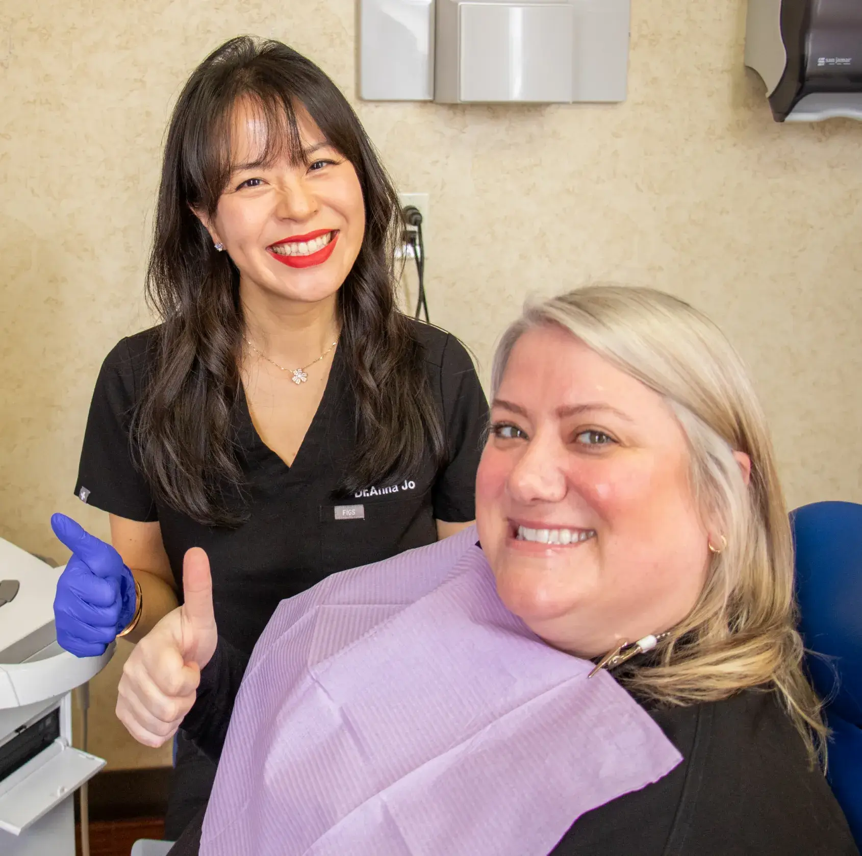 A dentist gives a thumbs up next to a smiling patient in a dental chair.
