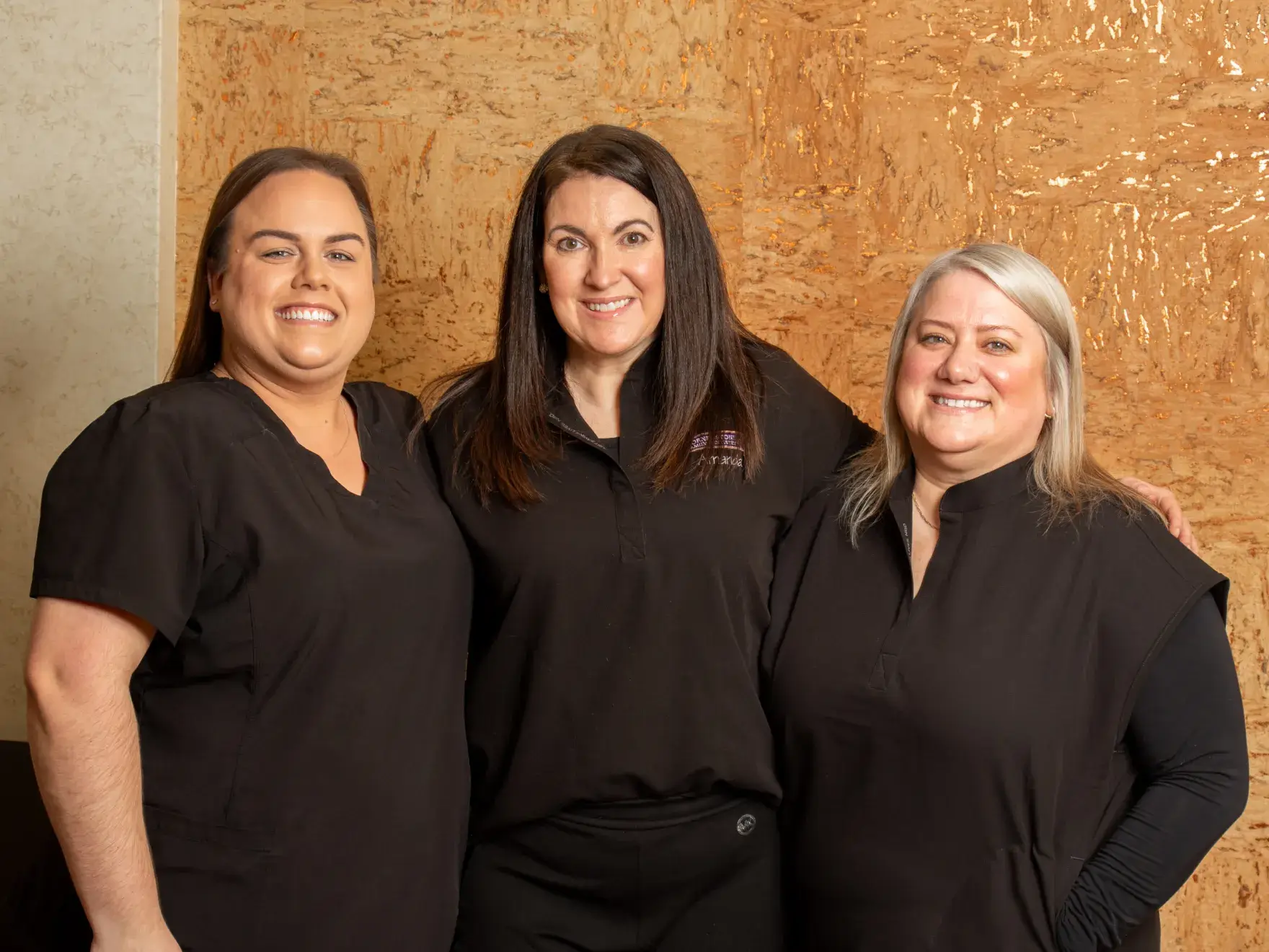 Three women in black clothing stand together smiling in front of a textured cork wall.