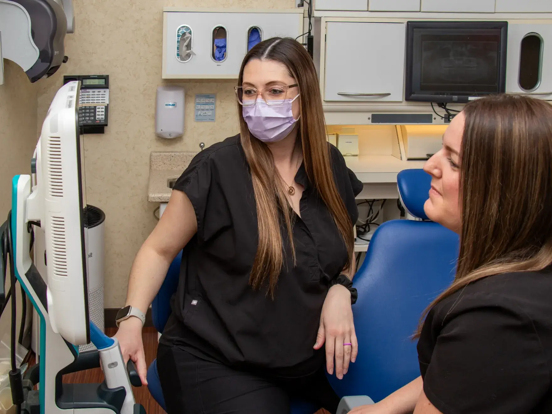 Two dental professionals in black scrubs discuss dental images on a computer screen.