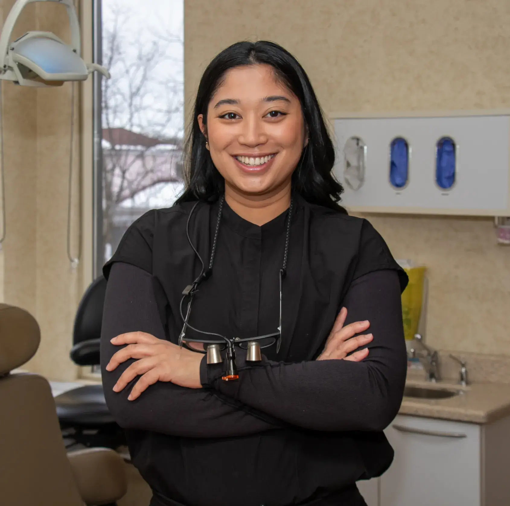 Smiling woman in a dental office, wearing a black uniform and dental magnifying glasses.