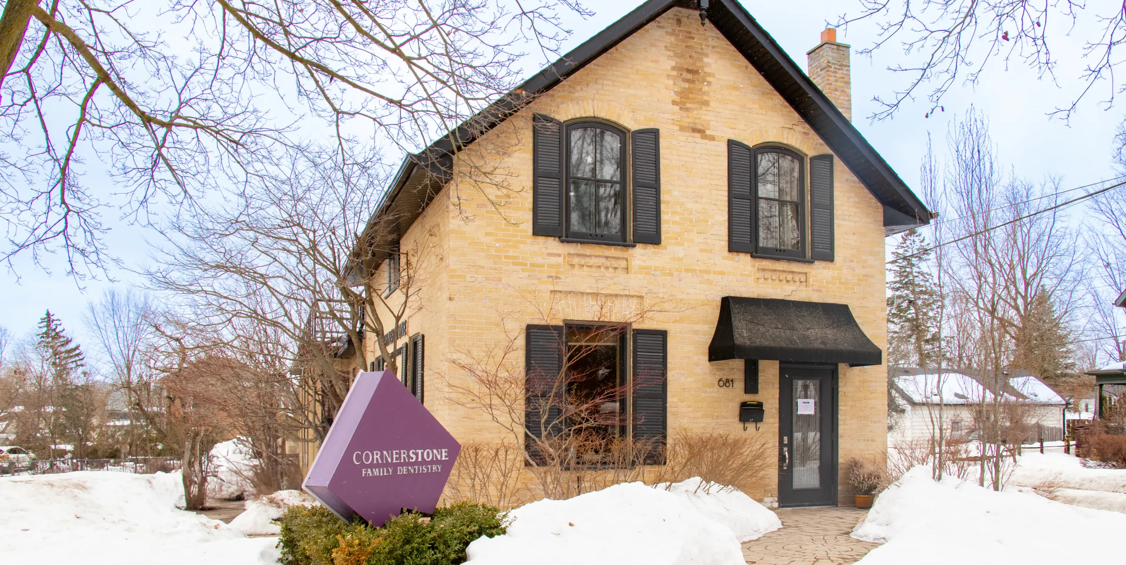 Two-story brick building with black shutters, surrounded by snow, featuring a large purple sign in front.