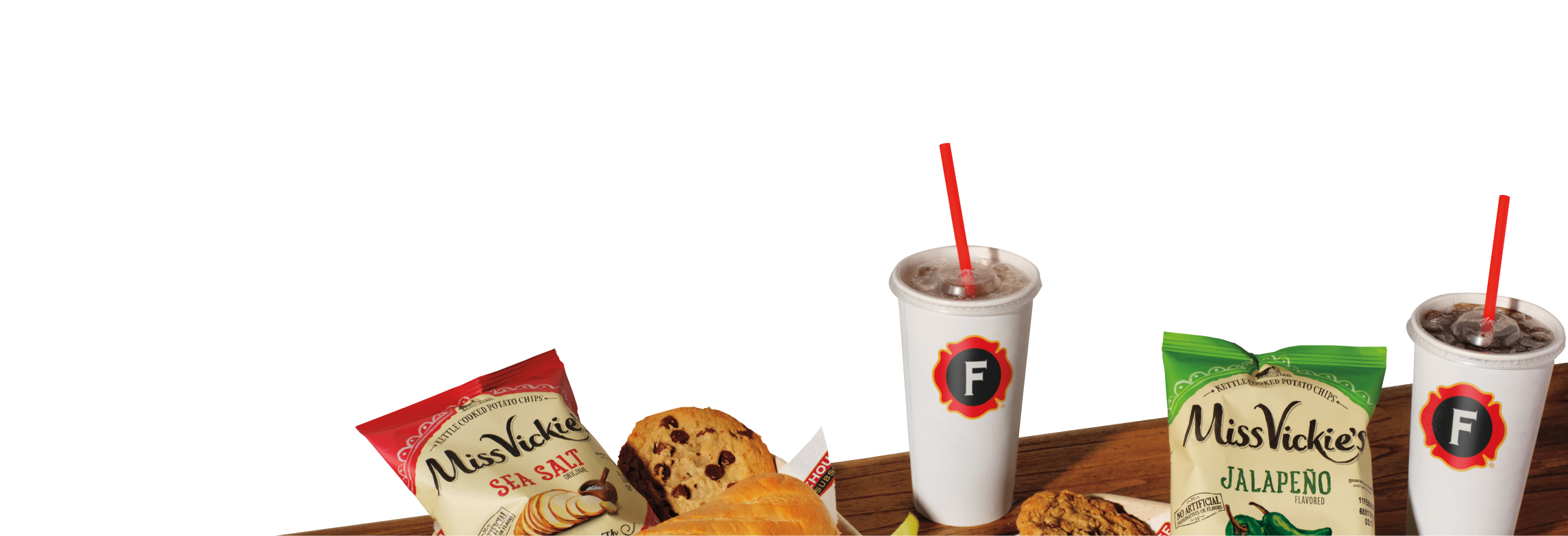 Table with two cups of soda with red straws and two bags of Miss Vickie's kettle-cooked potato chips, one Sea Salt and one Jalapeño flavor, alongside cookies and a sandwich.