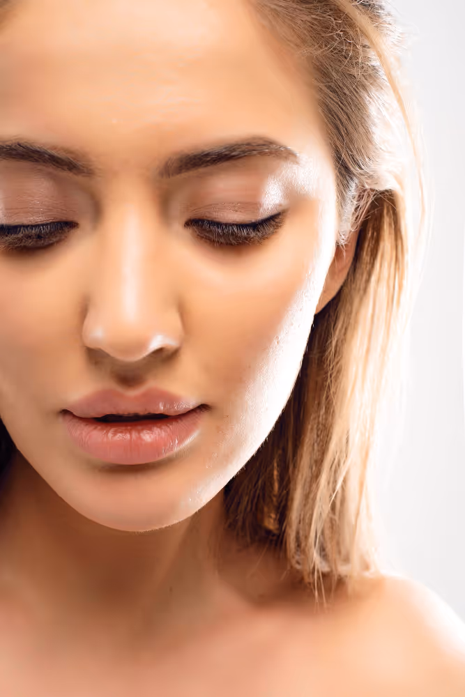 Nahaufnahme eines jungen Frauengesichts mit geschlossenen Augen und makelloser Haut vor hellem Hintergrund.