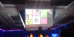 Large screen displaying colorful icons and images above a stage with people seated below in a dimly lit room.