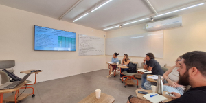 Group of six people seated in a classroom, watching a presentation on a large screen with notes on a whiteboard.