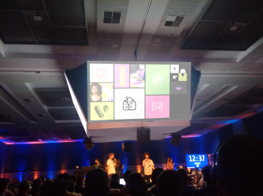Indoor event with a stage, speakers, large screen showing icons, and an audience seated under blue and purple lighting.