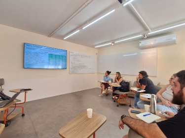Group of five people sitting in a classroom, attentively watching a screen with a spreadsheet and notes on a whiteboard.