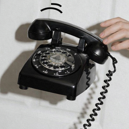 Hand lifting a black vintage rotary dial telephone with a coiled cord against a light background.