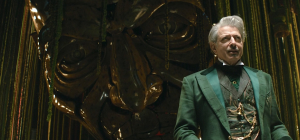 Homem mais velho vestindo terno verde e colete com relógio de bolso, em frente a grande estrutura escura ornamentada.