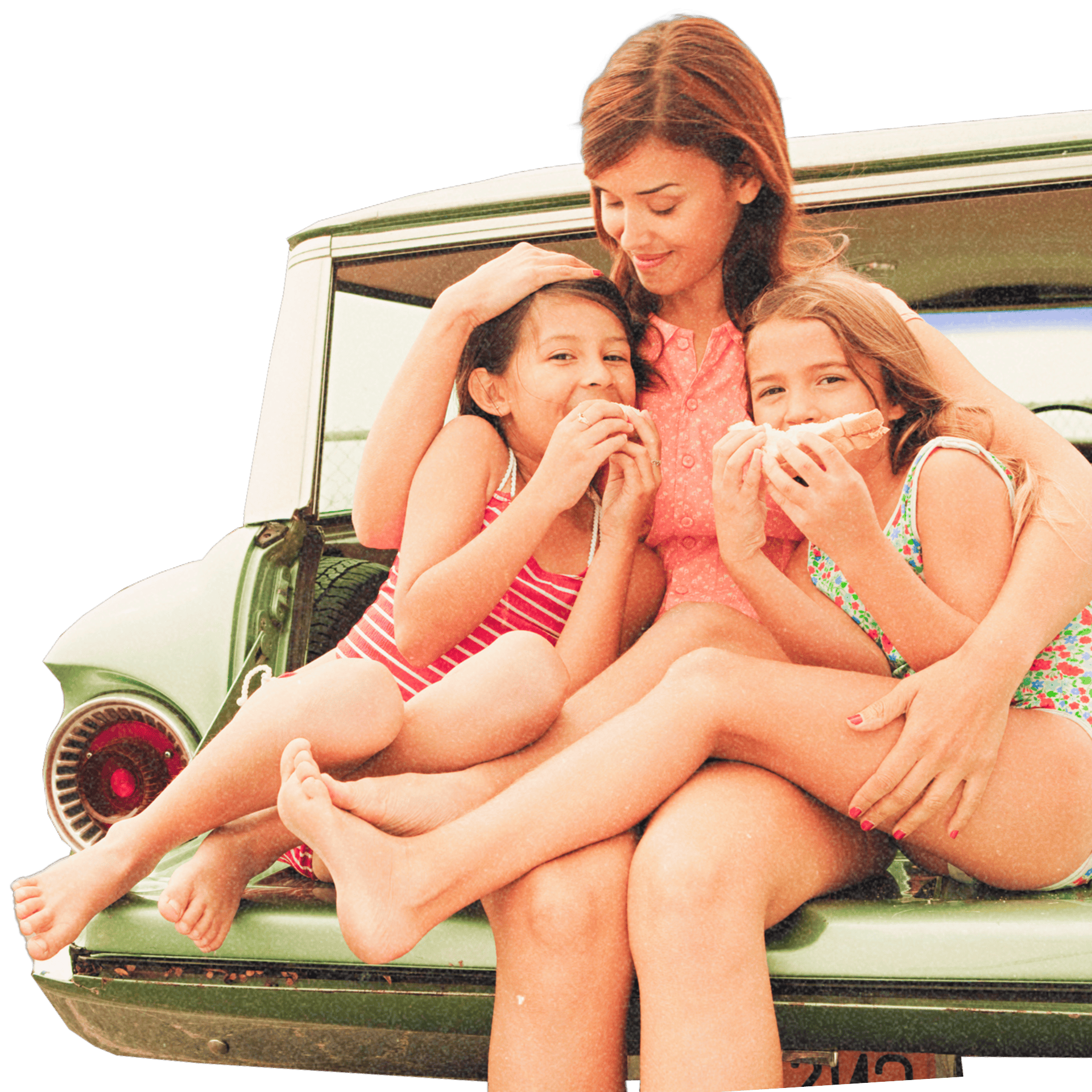 A mother embracing her two young daughters sitting on the open trunk of a green vintage car while the girls eat sandwiches.