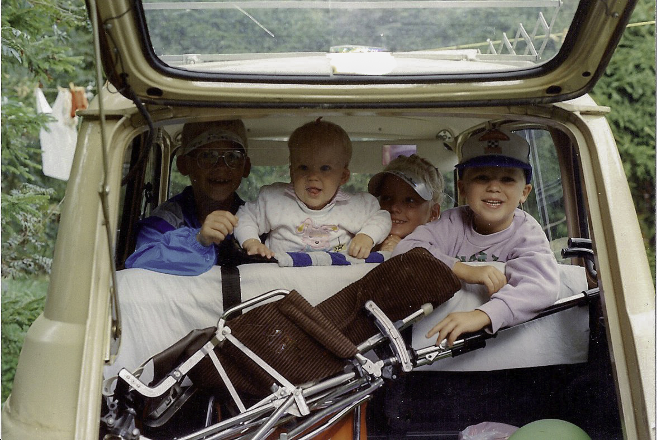 Four children sitting in the back of a car with the trunk open, including a toddler in a white sweater and three older kids, with a folded stroller in front of them.